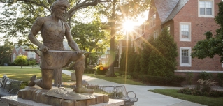 St. Ignatius statue on campus with sun shining through trees over building